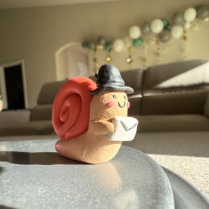 May include: A whimsical, handmade snail figurine with a red shell, wearing a black hat, and holding a white envelope. The snail has a smiling face with rosy cheeks. The figurine is likely made of clay or a similar material.