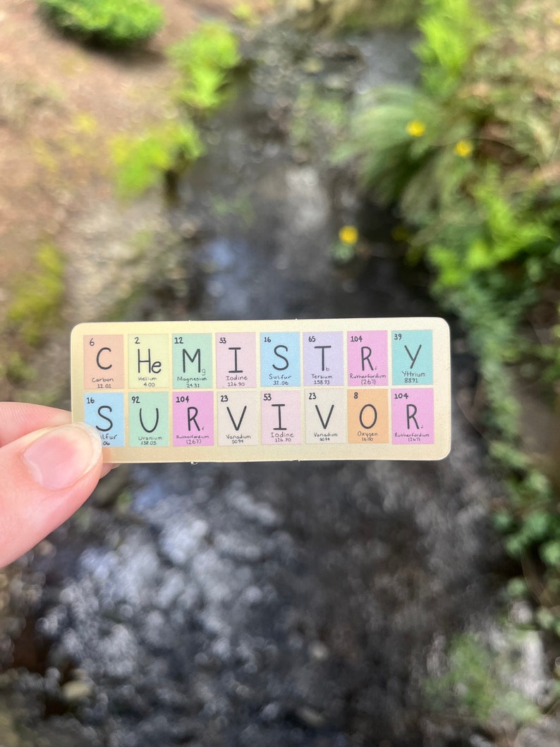 May include: A close-up of a white card with the word "CHEMISTRY" printed in black letters. Below the word "CHEMISTRY" is the word "SURVIVOR" spelled out using the chemical symbols for the elements. The card is being held by a hand.