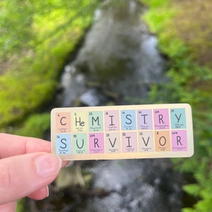May include: A colorful sticker with the word "SURVIVOR" spelled out using the chemical symbols for the elements. The sticker is held in a hand against a blurred background of green foliage and a stream of water.
