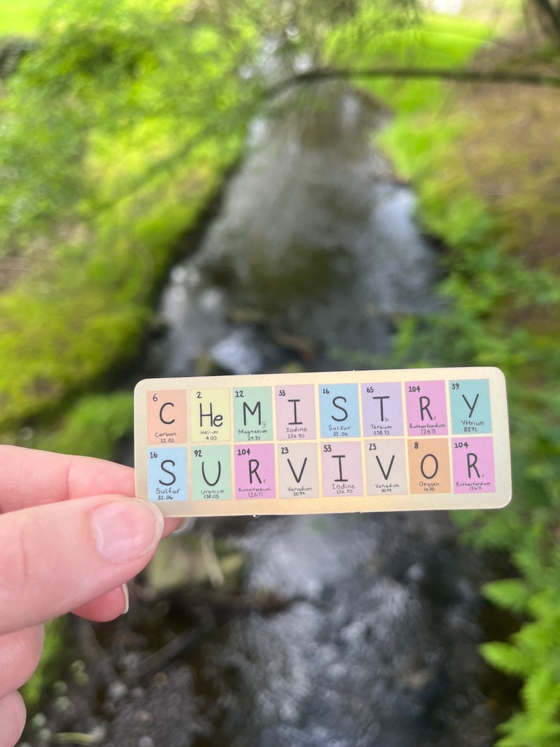 May include: A colorful sticker with the word "SURVIVOR" spelled out using the chemical symbols for the elements. The sticker is held in a hand against a blurred background of green foliage and a stream of water.