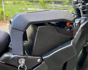 May include: Close-up of a black electric bicycle frame with a battery compartment and seat. The bike has a key in the ignition and suspension components. The background shows a green lawn and a fence.