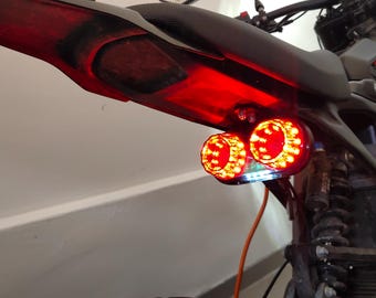 May include: A motorcycle's rear section with a red taillight and two round, bright red LED lights. The motorcycle is gray and black, with an orange wire visible. The lights are illuminated, suggesting they are in use.