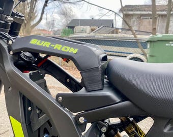 May include: Close-up of a black and gray electric motorcycle. The frame features the text "SUR-RON" in neon green. The seat is black and textured. The suspension and other mechanical components are visible.