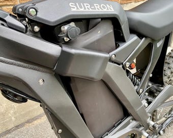 May include: Close-up of a black Sur-Ron electric dirt bike. The bike features a black frame, seat, and body panels. The name "SUR-RON" is visible on the top of the frame. The bike has a suspension system and a rear tire with a knobby tread.