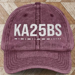 May include: A maroon baseball cap with the text "KA25BS" in white block letters. The hat has a curved brim and a distressed, vintage look. The text is above a dashed line.