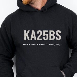 May include: Black hooded sweatshirt with a front pocket and the text "KA25BS" in a light grey font. Below the text is a line of dashes and dots. The word "BLACK" is printed in the upper right corner.