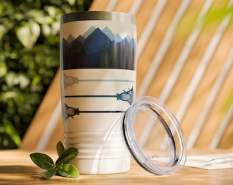 May include: A white stainless steel travel mug with a blue and white mountain scene and a lacrosse stick design. The mug has a clear lid.