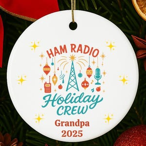 May include: White ceramic holiday ornament with the text "HAM RADIO Holiday CREW Grandpa 2025". The ornament features a design with a radio tower, microphone, and other radio-related icons. The ornament is surrounded by Christmas decorations.