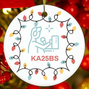 May include: White ceramic Christmas ornament with a festive design. The ornament features a string of colorful lights around the edge and a central graphic of a person with headphones and a microphone. The text "KA25BS" is below the graphic.