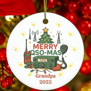 May include: White ceramic Christmas ornament with the text "MERRY QSO-MAS Grandpa 2025". The ornament features a Christmas tree, radio equipment, and a stocking. The ornament is hanging from a gold string.