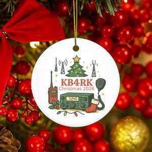 May include: A round white ceramic ornament featuring a green Christmas tree, radio towers, a handheld radio, a base station radio displaying 7.245, a red stocking, and the text KB4RK Christmas 2026, suspended by a gold string.