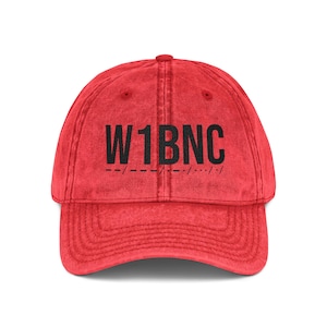 May include: A red baseball cap with a distressed, washed-out texture. The front features the black text W1BNC printed in a bold, sans-serif font, positioned directly above a line of black Morse code symbols consisting of dashes and dots.