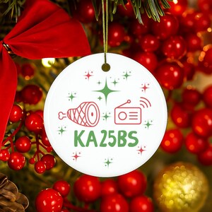 May include: A round white ceramic ornament featuring a red graphic of a ham, a red radio icon, and the green text KA25BS. The design is accented with small red and green starburst shapes, and the ornament hangs from a thin gold string.