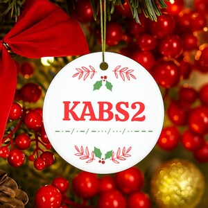 May include: A round white ceramic ornament featuring the red text KABS2 in the center, flanked by green holly leaves and red sprigs above and below. A green dashed line with dots sits below the text. It hangs from a gold string loop.