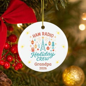 May include: A round white ceramic ornament featuring a colorful graphic of a radio tower, microphone, and handheld radio surrounded by holiday ornaments and stars. The text reads HAM RADIO Holiday CREW Grandpa 2026 in red and teal lettering.
