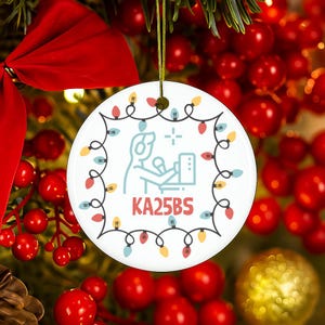 May include: A round white ceramic ornament featuring a light blue graphic of a person wearing headphones at a computer, with the text KA25BS below. The design is framed by a string of colorful Christmas lights in red, yellow, and blue hues.