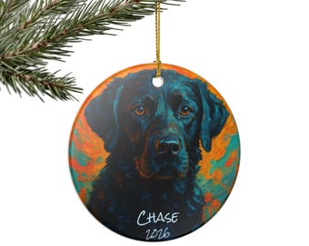 May include: A round ceramic ornament featuring a painted portrait of a black Labrador retriever against a vibrant orange and teal background. The name Chase and the year 2026 are printed in white script at the bottom. A gold string is attached.