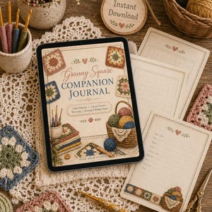 May include: A digital tablet displaying a Granny Square Companion Journal cover with crochet motifs, alongside printed journal pages, a crochet hook, scissors, and yarn balls. The journal cover features text, a basket of yarn, and granny squares.