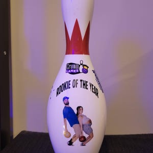 May include: A white bowling pin featuring a red neck stripe, the Crown Lanes logo, and the text "ROOKIE OF THE YEAR" above a graphic of two people holding bowling balls. The year "2025" is printed in black at the bottom of the bowling pin.