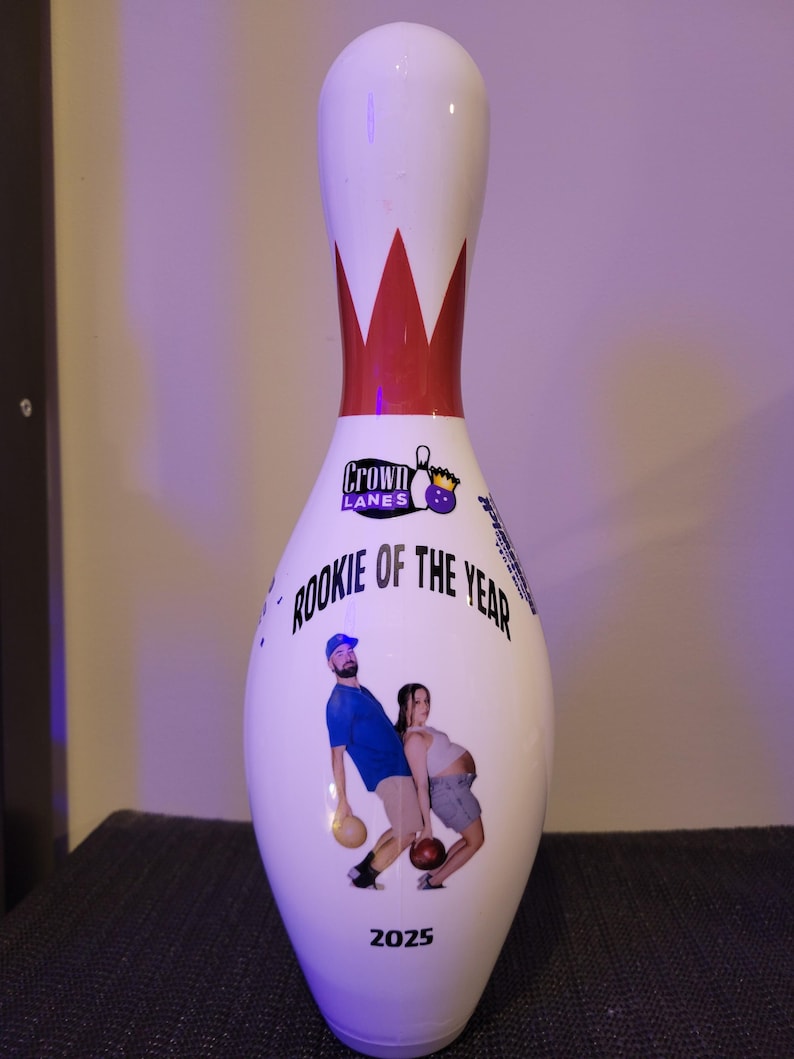 May include: A white bowling pin featuring a red neck stripe, the Crown Lanes logo, and the text "ROOKIE OF THE YEAR" above a graphic of two people holding bowling balls. The year "2025" is printed in black at the bottom of the bowling pin.