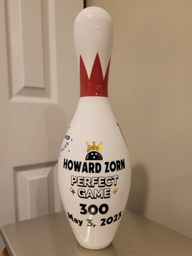 May include: A white bowling pin featuring a red crown graphic at the neck and a gold crown icon above the text HOWARD ZORN PERFECT GAME 300 May 3, 2023. The pin has a smooth, glossy finish and includes small gold star accents near the text.