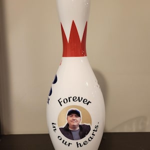 May include: A white bowling pin featuring a red crown-like graphic at the neck. The body displays a circular photo of a person wearing a cap, surrounded by the black text "Forever in our hearts." A partial blue logo is visible on the side.