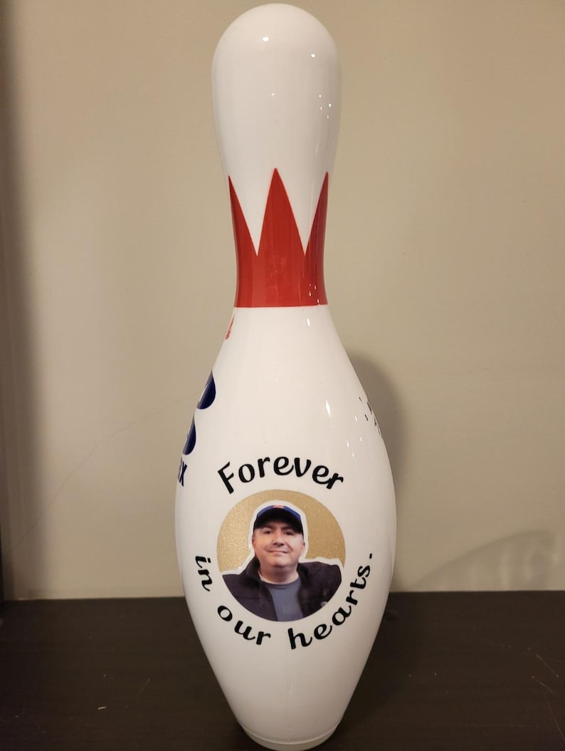 May include: A white bowling pin featuring a red crown-like graphic at the neck. The body displays a circular photo of a person wearing a cap, surrounded by the black text "Forever in our hearts." A partial blue logo is visible on the side.