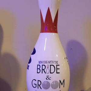 May include: A white bowling pin featuring a red neck stripe and black text that reads BOWLING WITH THE BRIDE & GROOM THE BLEICHERS 9.28.25. The letter I in BRIDE is a small bowling pin, and the two Os in GROOM are textured silver bowling balls.