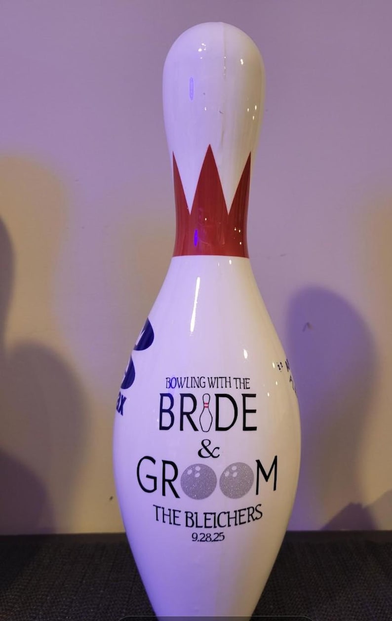 May include: A white bowling pin featuring a red neck stripe and black text that reads BOWLING WITH THE BRIDE & GROOM THE BLEICHERS 9.28.25. The letter I in BRIDE is a small bowling pin, and the two Os in GROOM are textured silver bowling balls.