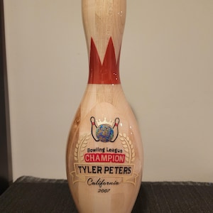 May include: A light-colored wooden bowling pin featuring a red crown graphic at the neck. The body displays a colorful globe icon, the text Bowling League CHAMPION, the name TYLER PETERS, the location California, and the year 2007 in black lettering.