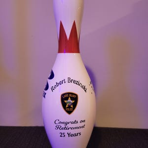 May include: A white bowling pin featuring a red crown graphic at the neck. The body displays the text Robert Brezinski, a Lake Bluff Police badge emblem, and the text Congrats on Retirement 25 Years printed in black ink on the front surface.