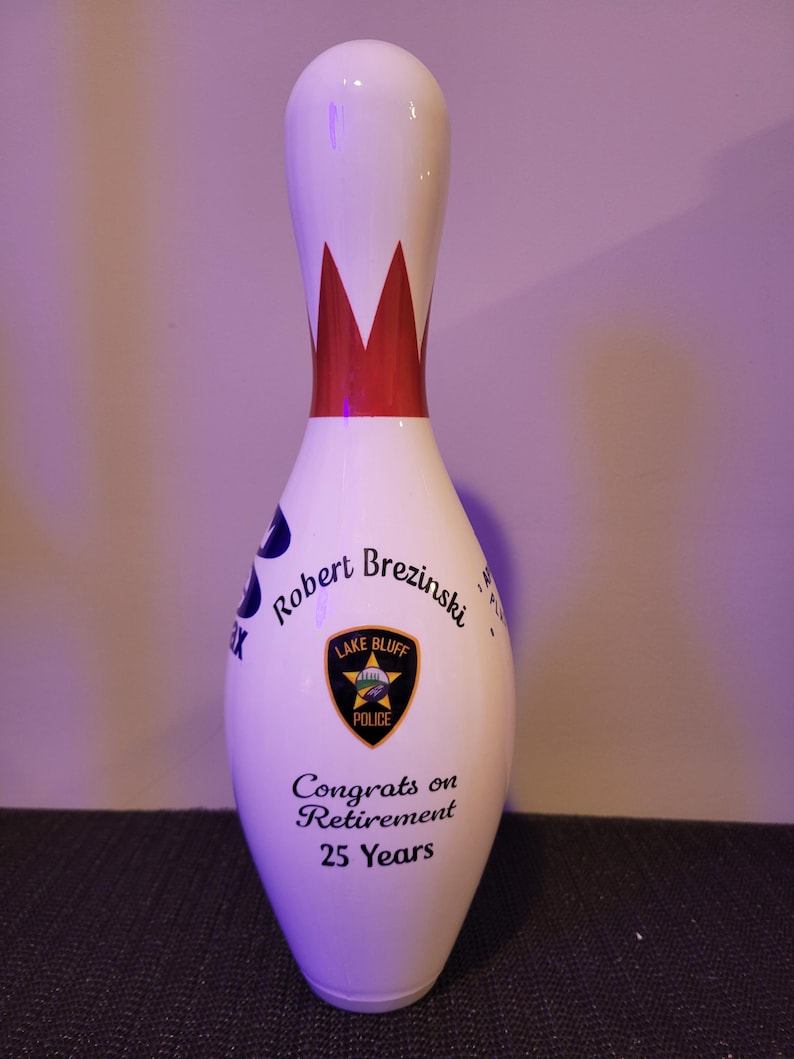May include: A white bowling pin featuring a red crown graphic at the neck. The body displays the text Robert Brezinski, a Lake Bluff Police badge emblem, and the text Congrats on Retirement 25 Years printed in black ink on the front surface.