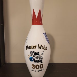 May include: A white bowling pin with a red and white top. The pin is decorated with the text "Master Webb King of the Lanes 300 4-1-2024" and a bowling ball and pin graphic. A Brunswick logo is visible.
