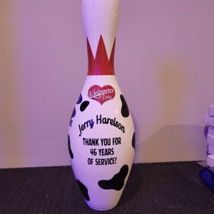 May include: A white bowling pin featuring a black cow-print pattern, a red crown design at the neck, and a red heart logo labeled Kleinpeter Dairy. The text reads: Jerry Harelson THANK YOU FOR 46 YEARS OF SERVICE! The pin has a smooth, glossy finish.