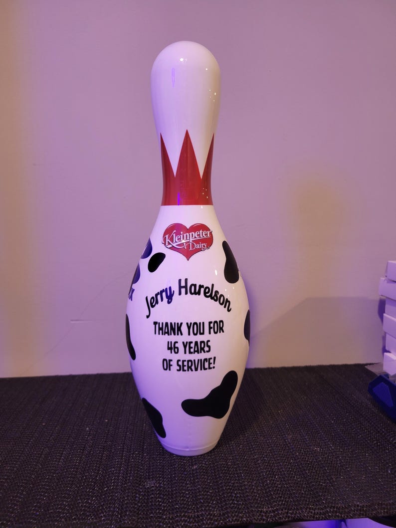 May include: A white bowling pin featuring a black cow-print pattern, a red crown design at the neck, and a red heart logo labeled Kleinpeter Dairy. The text reads: Jerry Harelson THANK YOU FOR 46 YEARS OF SERVICE! The pin has a smooth, glossy finish.