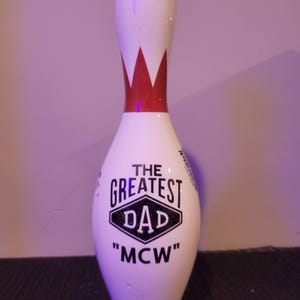 May include: A white bowling pin featuring a red crown graphic at the neck and black text on the body that reads THE GREATEST DAD and MCW inside a diamond shape. The surface is smooth and glossy, with a rounded top and a tapered base design.