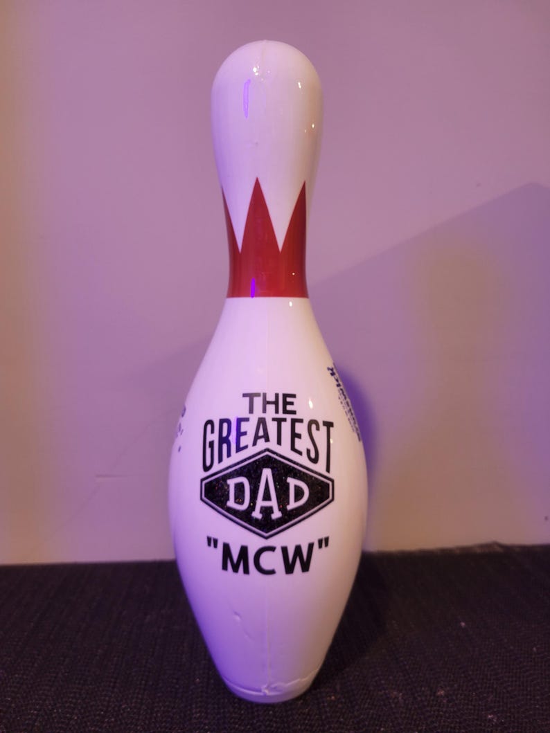 May include: A white bowling pin featuring a red crown graphic at the neck and black text on the body that reads THE GREATEST DAD and MCW inside a diamond shape. The surface is smooth and glossy, with a rounded top and a tapered base design.