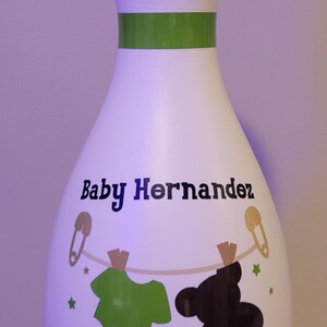 May include: A white bowling pin featuring a green neck stripe and the text Baby Hernandez in black. Below the text, a clothesline graphic displays a green onesie and a black teddy bear silhouette, accented by gold safety pins and small stars.