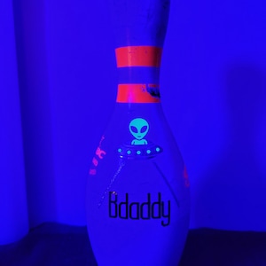 May include: A white bowling pin illuminated under black light, featuring a glowing green alien in a flying saucer graphic, two horizontal orange stripes on the neck, and the black text "Bdaddy" printed on the lower body of the pin.