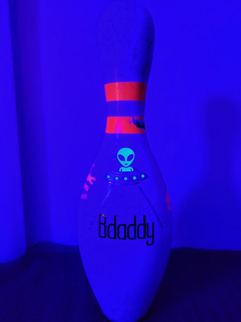May include: A white bowling pin illuminated under black light, featuring a glowing green alien in a flying saucer graphic, two horizontal orange stripes on the neck, and the black text "Bdaddy" printed on the lower body of the pin.