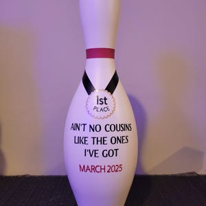 May include: A white bowling pin featuring a pink neck stripe and a black ribbon graphic with a gold 1st Place medal emblem. The text reads: AIN'T NO COUSINS LIKE THE ONES I'VE GOT and MARCH 2025 in black and red lettering on the front surface.