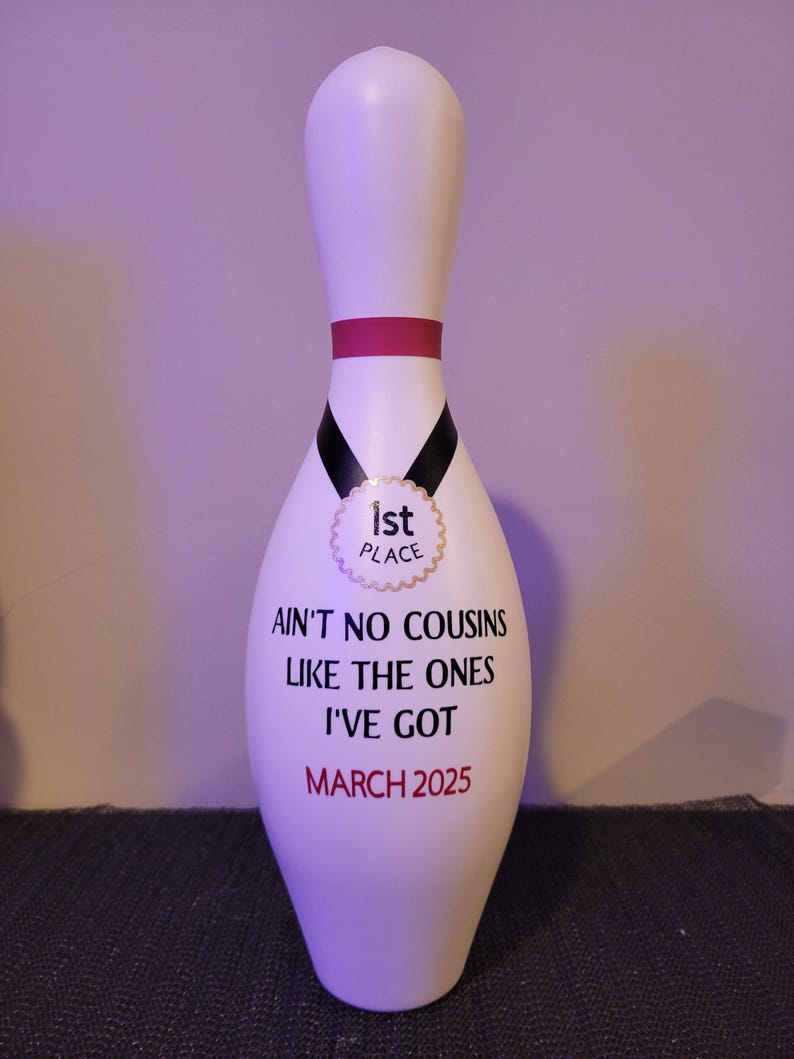 May include: A white bowling pin featuring a pink neck stripe and a black ribbon graphic with a gold 1st Place medal emblem. The text reads: AIN'T NO COUSINS LIKE THE ONES I'VE GOT and MARCH 2025 in black and red lettering on the front surface.