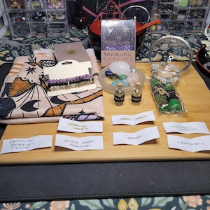 Puede incluir: Una colección de artículos para la práctica espiritual, que incluye un tapiz, cartas de oráculo, cartas de tarot, un brazalete de cuentas de cristal, una pila de brazaletes de protección, velas vestidas, hierbas y botellas de aceite. El texto en las cartas dice "Sanación chamánica" y "Cartas de tarot".