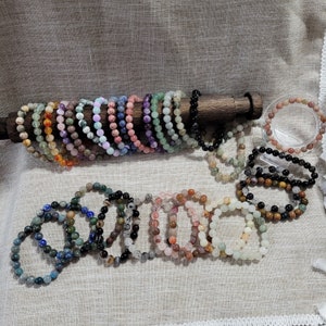 Peut inclure: Un ensemble de 20 bracelets en pierres précieuses de couleurs différentes, chacun avec un motif unique. Les bracelets sont faits de petites perles rondes et sont enfilés sur un cordon élastique. Ils sont présentés sur un support en bois.