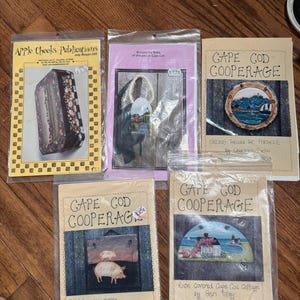 May include: A collection of Cape Cod Cooperage slate patterns and Apple Cheeks Publications patterns. The patterns feature coastal scenes, including a cottage, a lighthouse, and a pig. The patterns are in plastic sleeves.