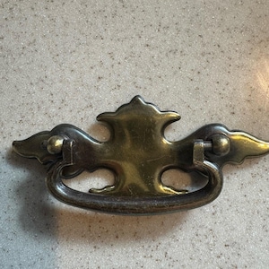 May include: A vintage-style brass drawer pull with a decorative design. The pull features a curved handle and ornate details, including stylized wings on either side. The hardware is likely for a cabinet or dresser.