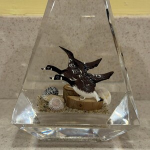 May include: A clear, geometric resin sculpture featuring three detailed duck figurines in flight. The ducks are brown and white, set against a backdrop of sand, seashells, and a small wooden log. The sculpture is a decorative item.