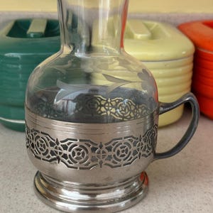 May include: A clear glass pitcher with a silver metal base and handle. The base has a decorative, cut-out design.