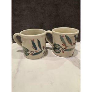 May include: Two off-white ceramic mugs featuring a hand-painted botanical design of green leaves and small red berries on thin brown stems. Each mug has a sturdy curved handle and a smooth, rounded rim, showcasing a consistent floral pattern.