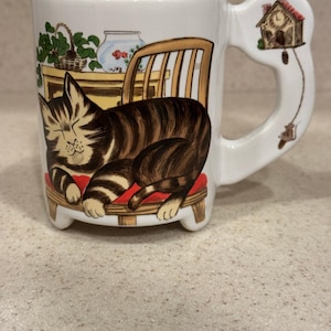 May include: White ceramic mug featuring a sleeping tabby cat on a wooden chair with a red cushion. The mug's handle is shaped with a decorative birdhouse design. The mug has a whimsical, illustrated style.